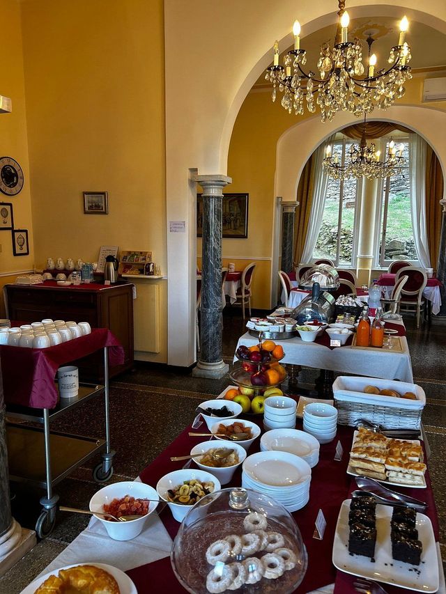 Colazione a buffet in una grande sala da pranzo, con vari piatti sui tavoli, un lampadario e porte ad arco.