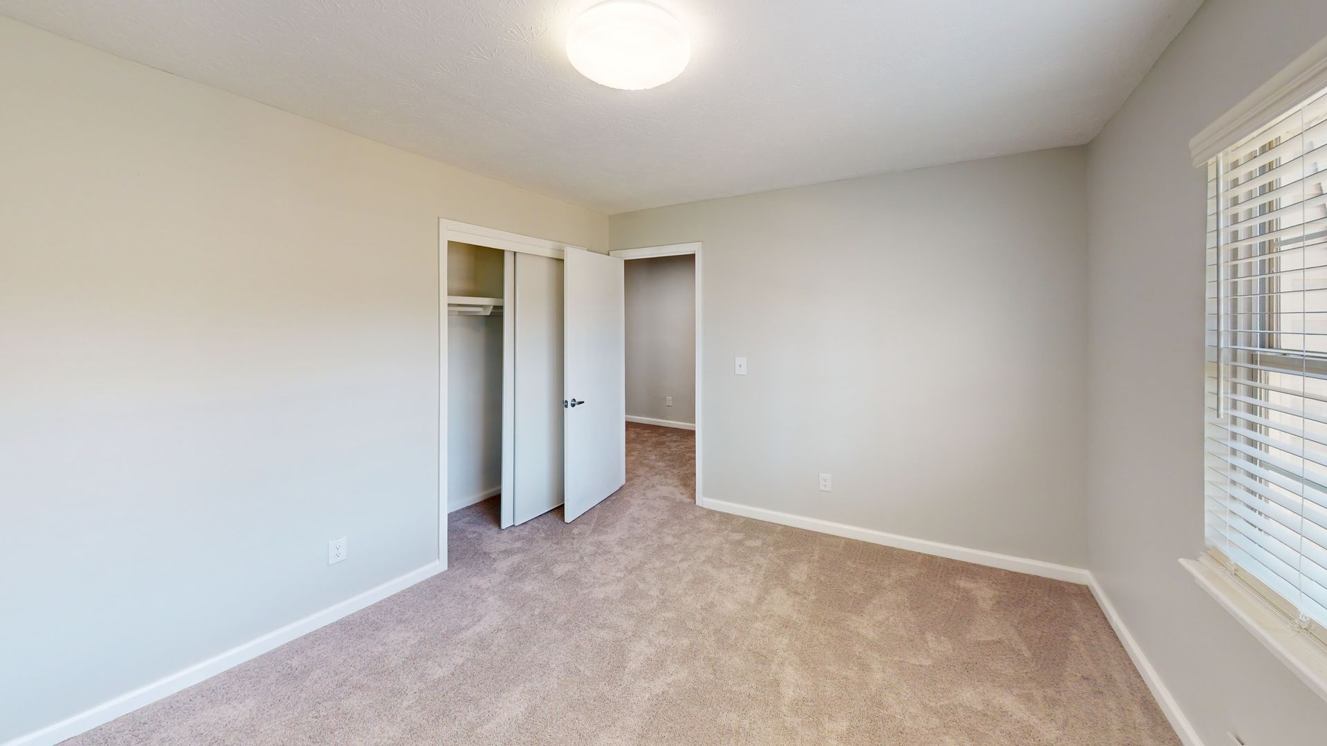 Empty beige bedroom with carpet, open closet, and window blinds letting in light.