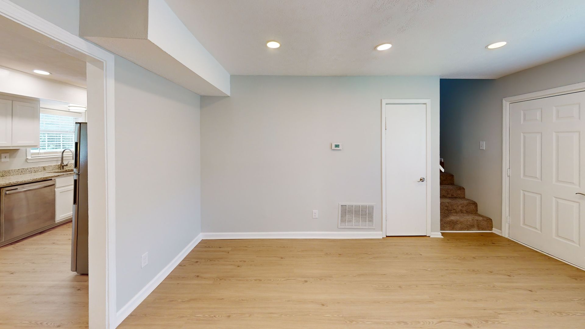 Empty modern living room with light wood floors, white walls, and stairs beside a closed door.