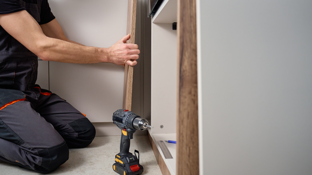 A worker kneels on the floor, using a power drill to install a wooden panel onto a kitchen cabinet.
