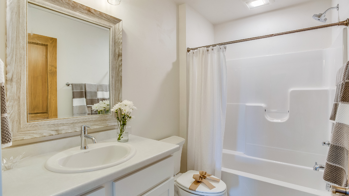 A bright bathroom with a light-framed vanity mirror, a white sink, a toilet, and a white shower-tub unit with a curtain.