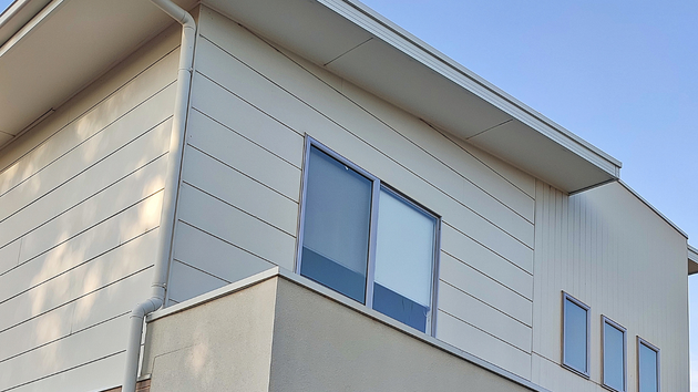 Low-angle view of a modern two-story building with white horizontal siding and a gray stucco balcony under a blue sky.