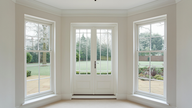 A set of white French doors flanked by two sash windows in a room with light-colored walls, overlooking a garden.