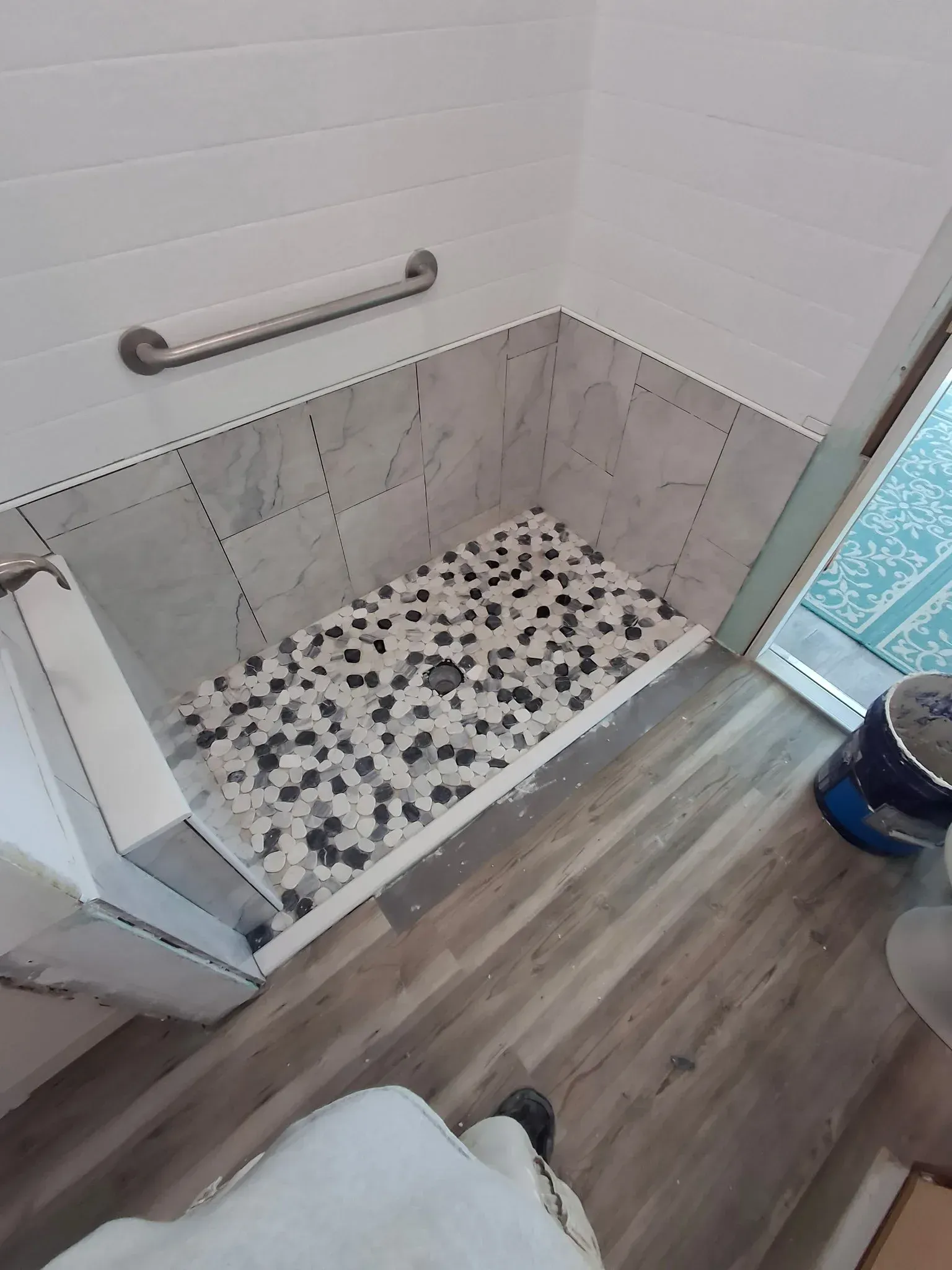 A walk-in shower with a pebble-stone floor, marble-look tiled walls, a metal grab bar, and a partial glass door.