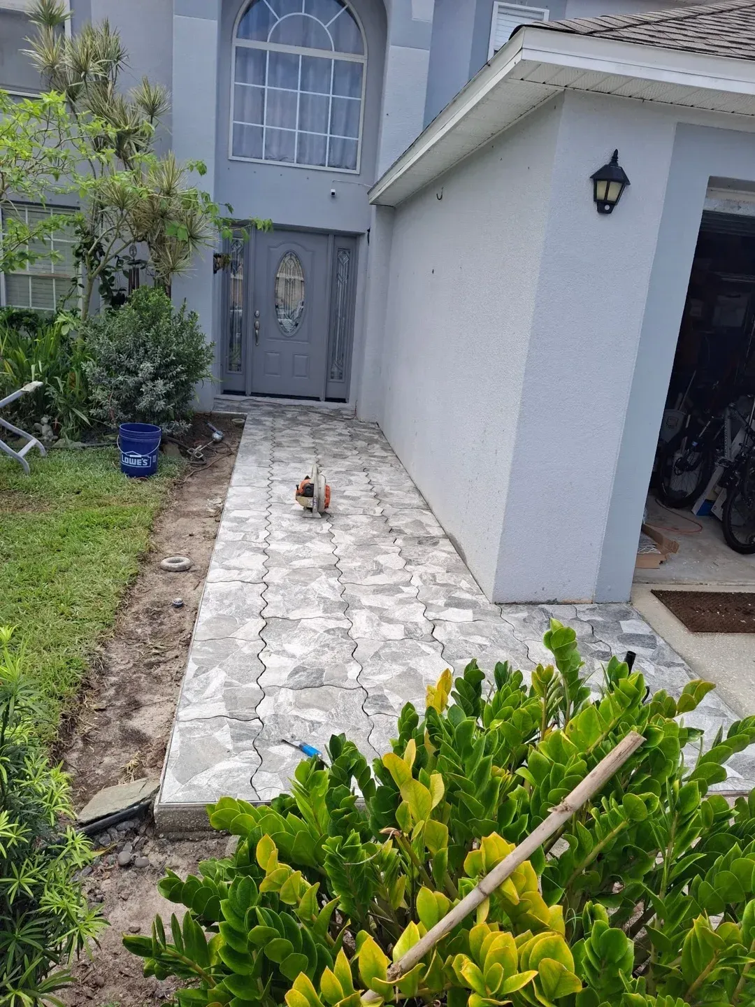 A gray tiled walkway leads from a manicured lawn to the front door of a suburban house, with a garage on the right.