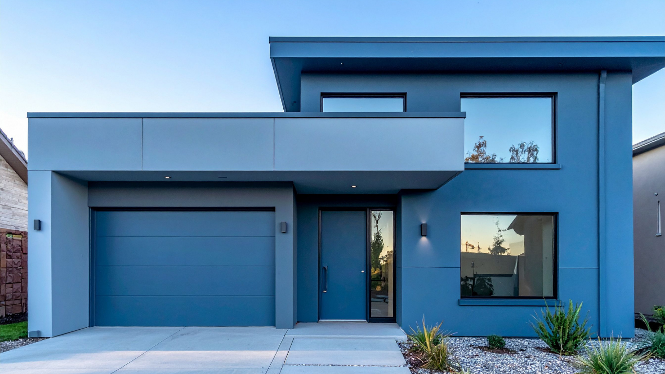 A modern two-story house with a flat roof, blue exterior walls, a double garage, and a light grey cantilevered section.
