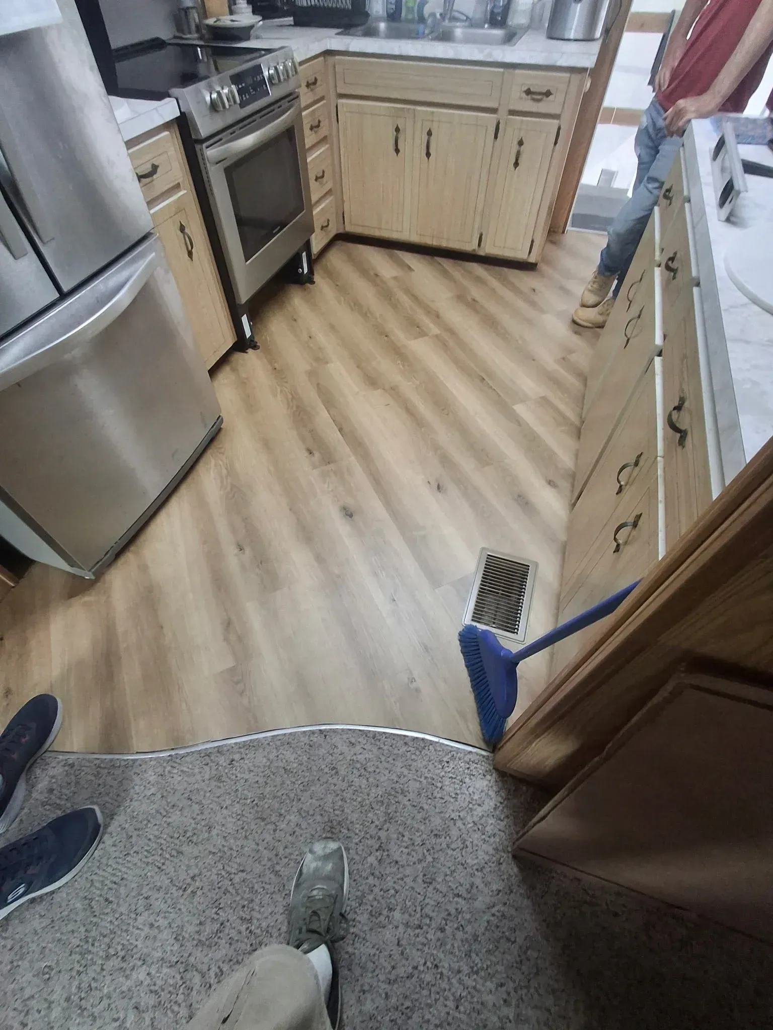A kitchen with light wood-tone flooring, stainless steel appliances, and wood cabinets, viewed from a carpeted area.