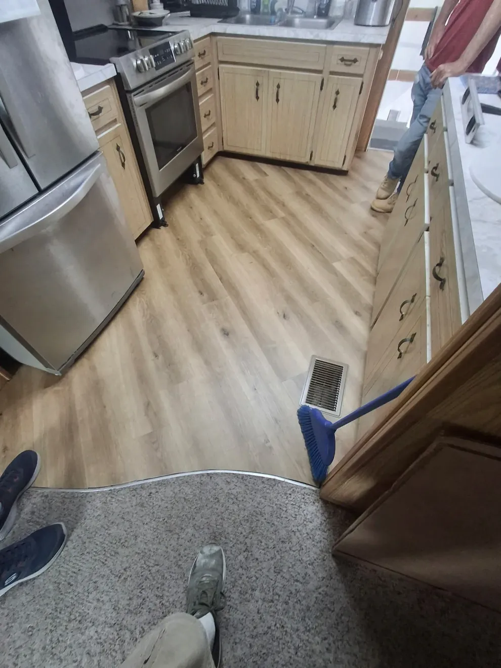 A first-person view of a kitchen with light wood-tone flooring, stainless steel appliances, and a blue broom.