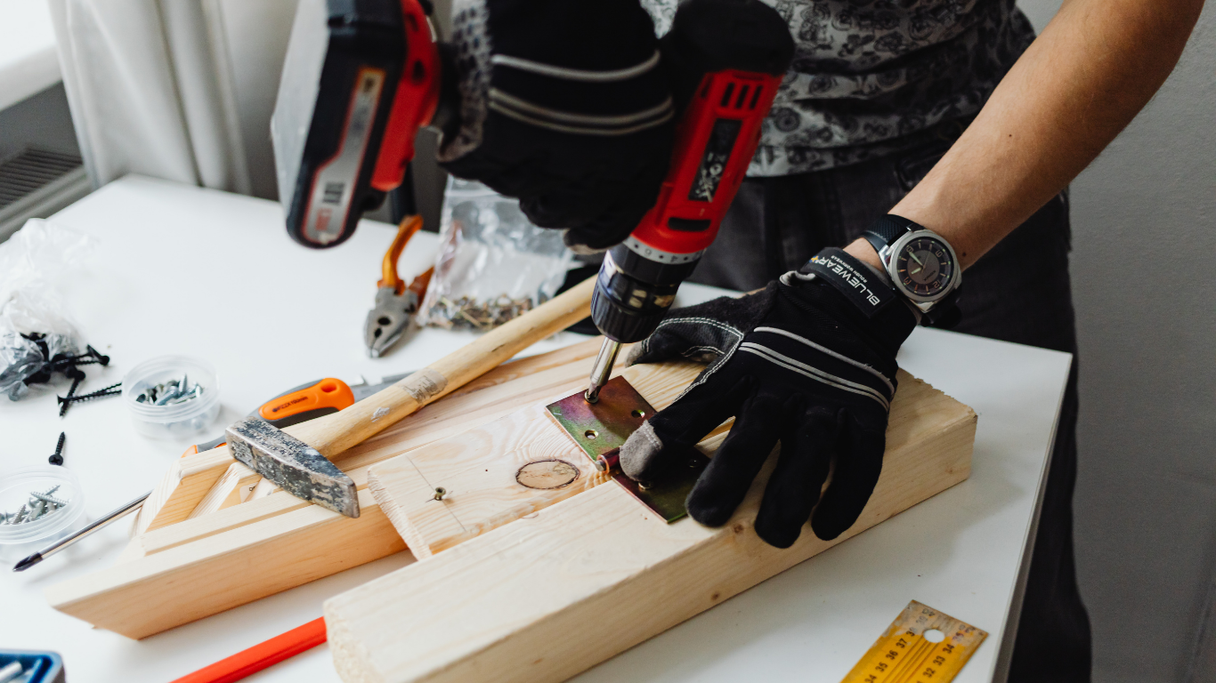 A person wearing black work gloves uses a red power drill to attach a metal hinge to a piece of wood on a white desk.