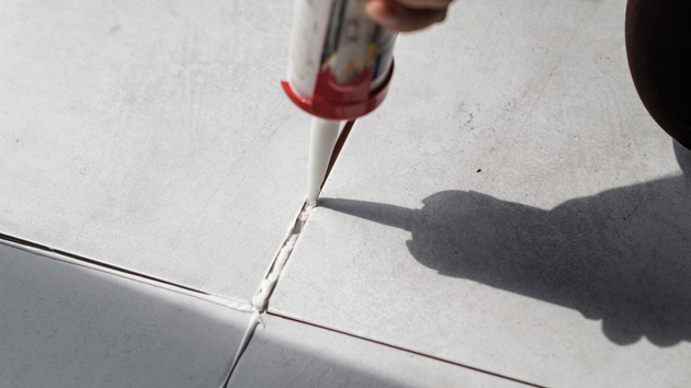 A hand holds a caulking gun to apply a bead of white sealant into the joint between two light gray floor tiles.