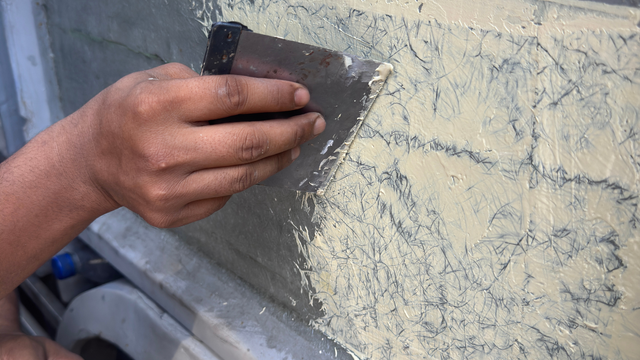 A hand uses a metal putty knife to spread light-colored, textured filler over a gray surface.