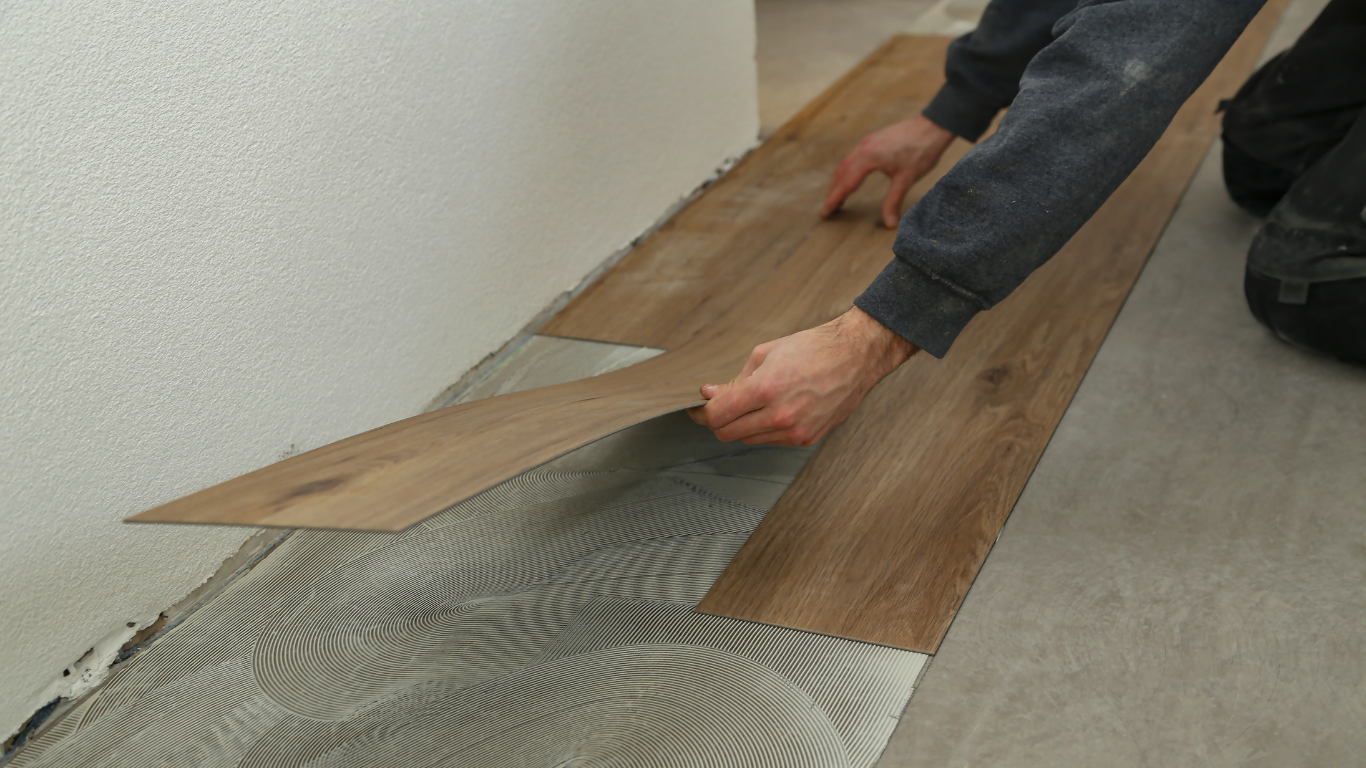 A person kneeling and installing wood-look vinyl planks onto a floor with spread adhesive.