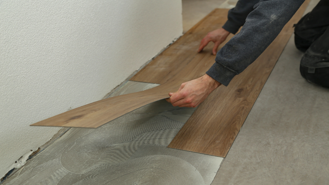 A person installing wood-look vinyl flooring over a thin layer of adhesive on a subfloor next to a white wall.
