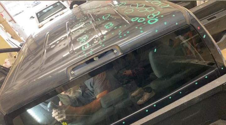 Person inside a vehicle's cab, likely repairing the windshield. Green markings, sunlight, grey vehicle roof and window.