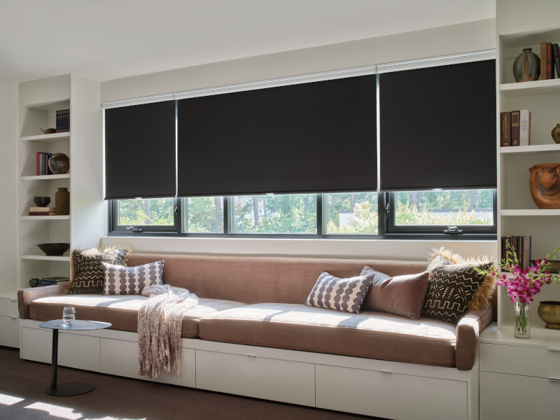Cozy window seat with dark roller shades, overlooking a window. Built-in shelving and neutral pillows.