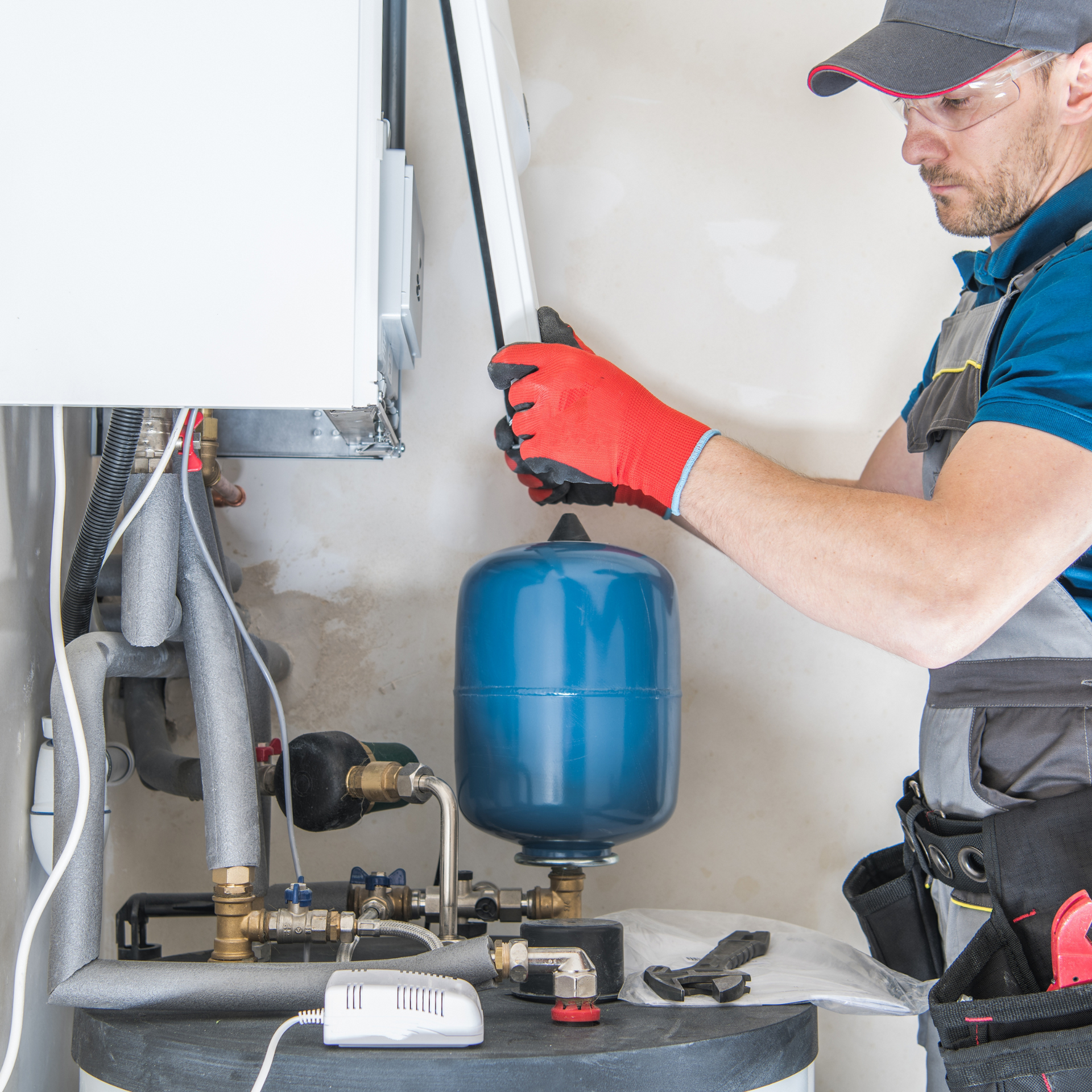 Plumber in gray hat, red gloves, repairing a water heater. Blue expansion tank visible.