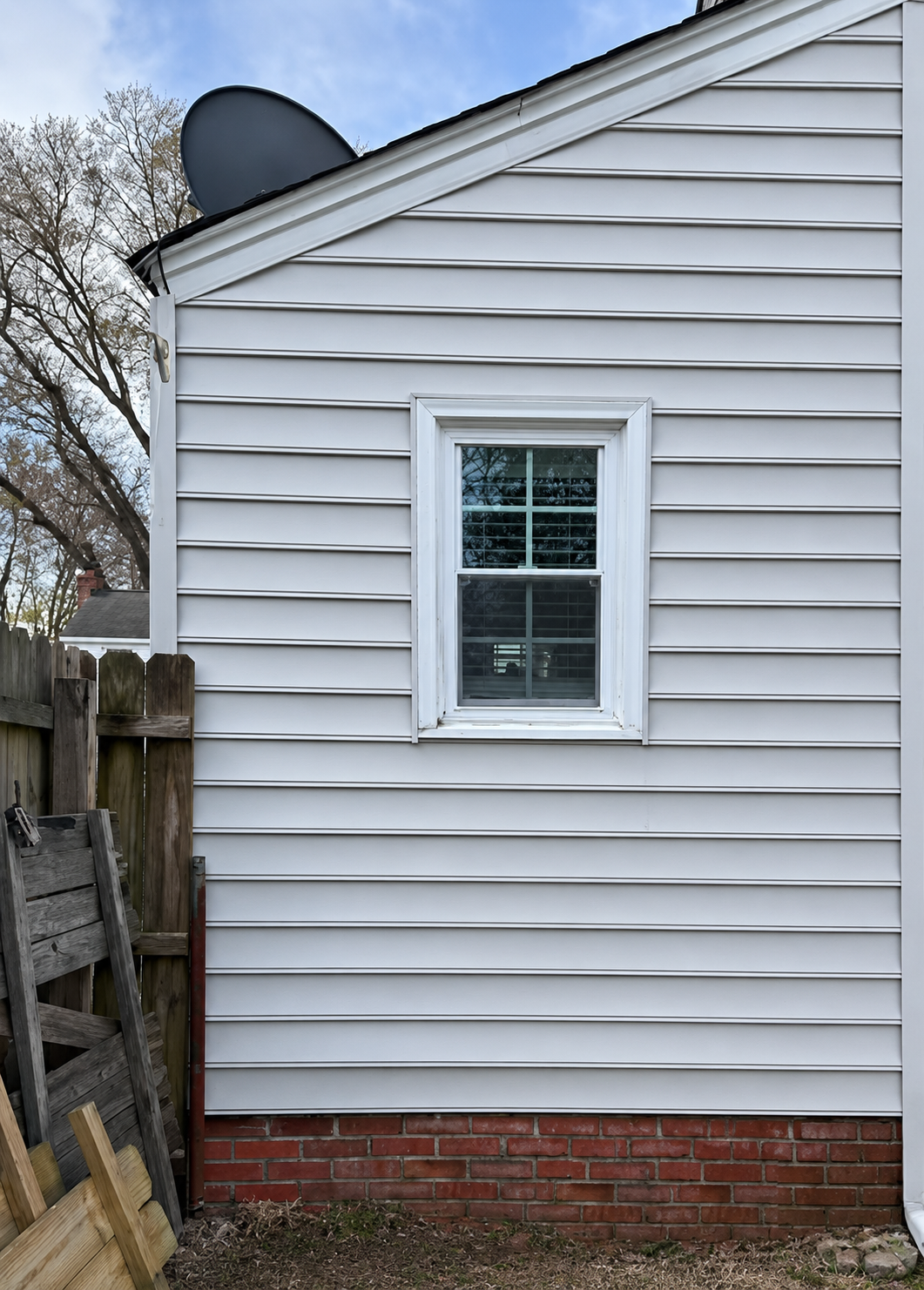 White vinyl house wall with a small window, brick foundation, and a fence along the left side.