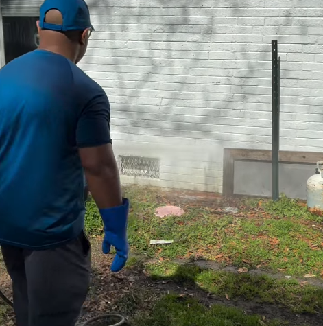 Worker in blue shirt and gloves near a white brick wall, inspecting the ground in a backyard.