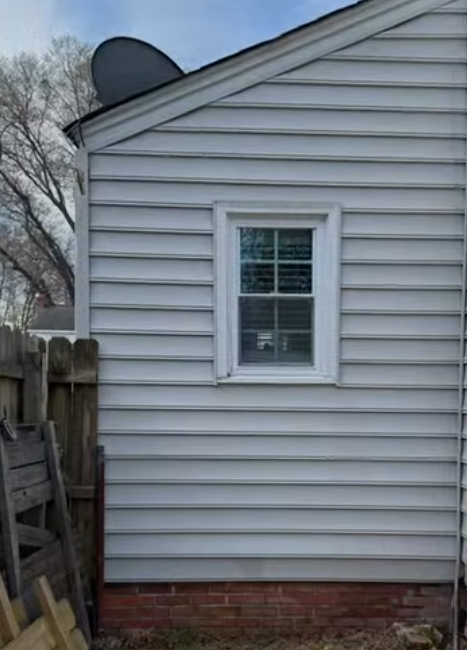 Side of a light gray house with a small white-framed window and brick foundation next to a wooden fence.