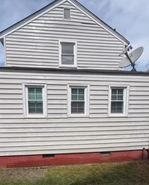 Side view of a white house with three windows, red foundation trim, and a satellite dish on the roof.