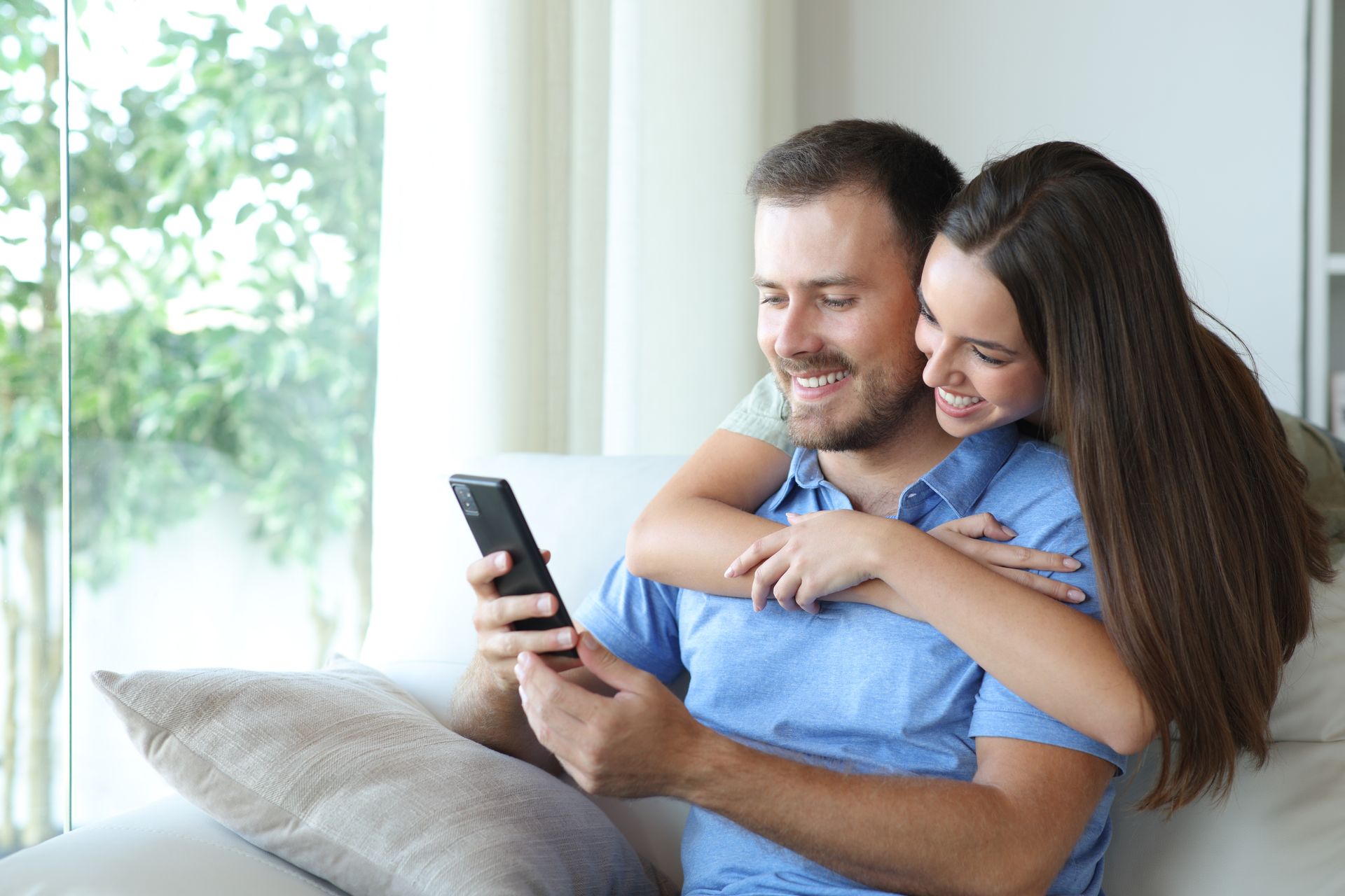 Couple smiling on a couch, cuddling and looking at a smartphone in a bright living room.