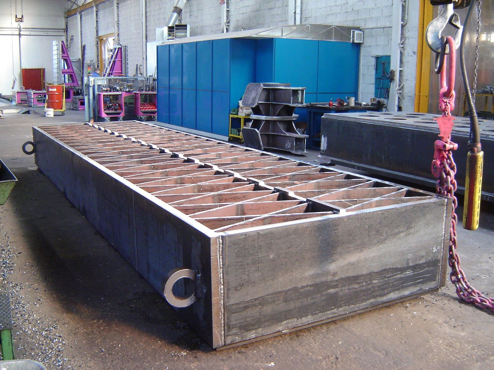 Officina industriale con componenti metallici e un operaio con indumenti protettivi arancioni che salda scintille blu