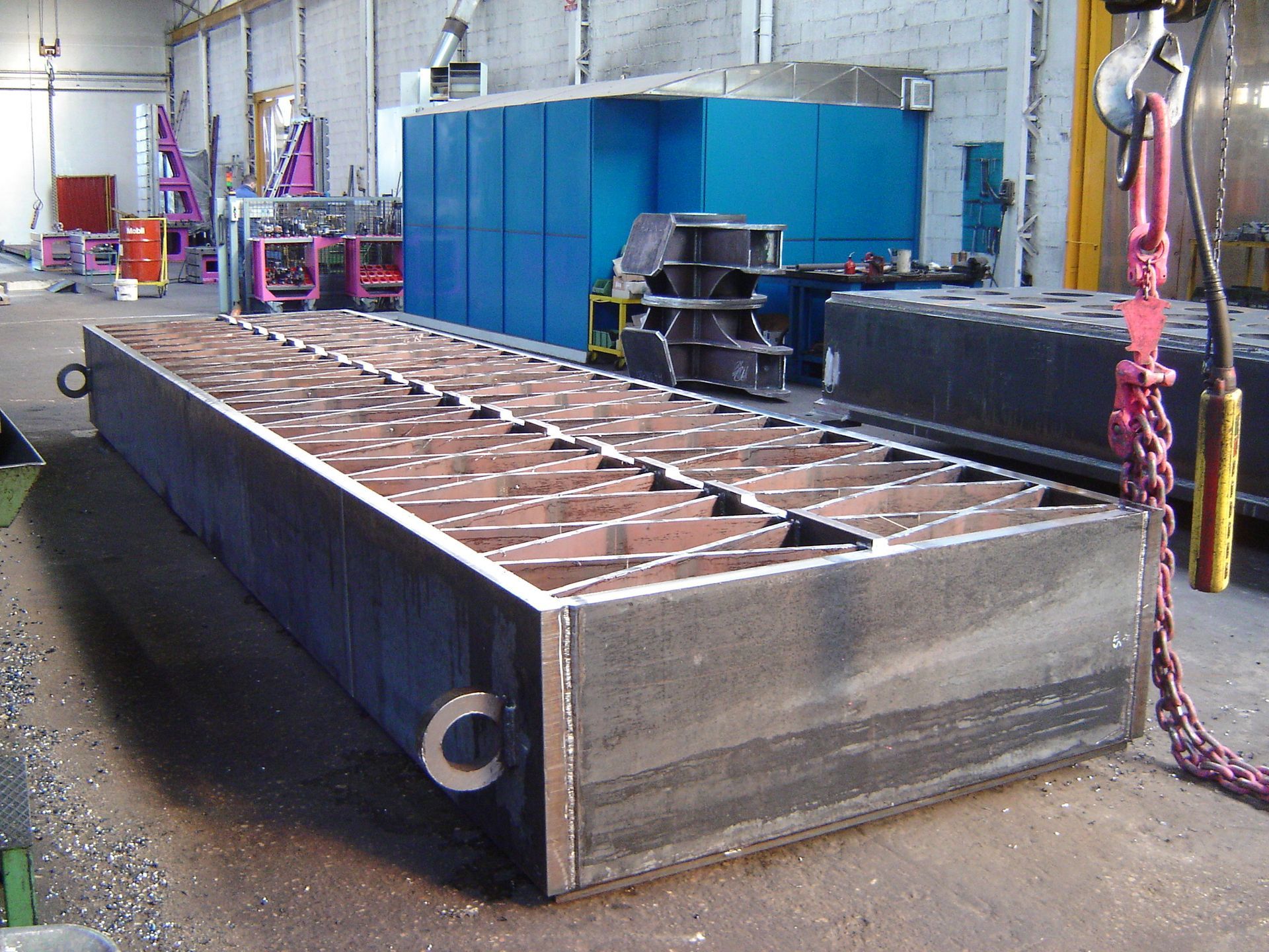 Operai che saldano parti metalliche in un'officina industriale poco illuminata, con grandi macchinari e telai nelle vicinanze.