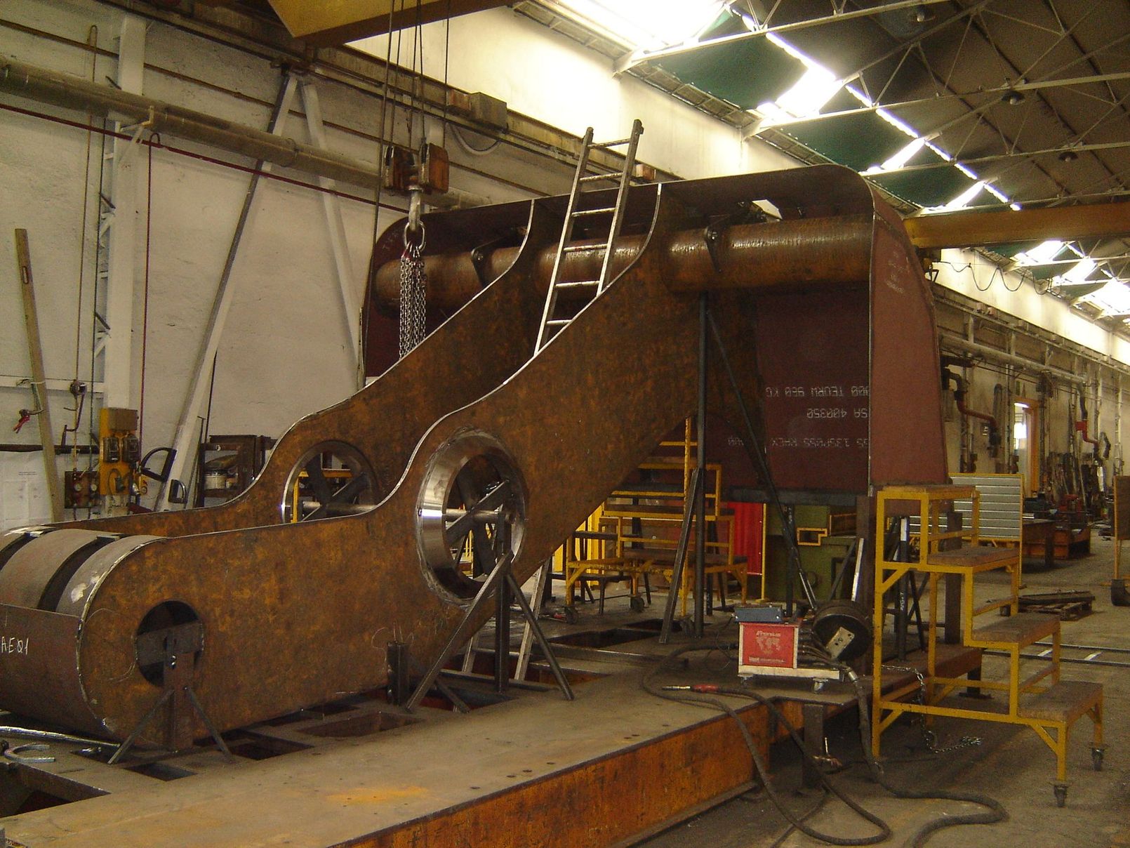 Officina industriale con ampi recinti metallici per macchinari, aperture circolari e una gru a portale blu all'interno.