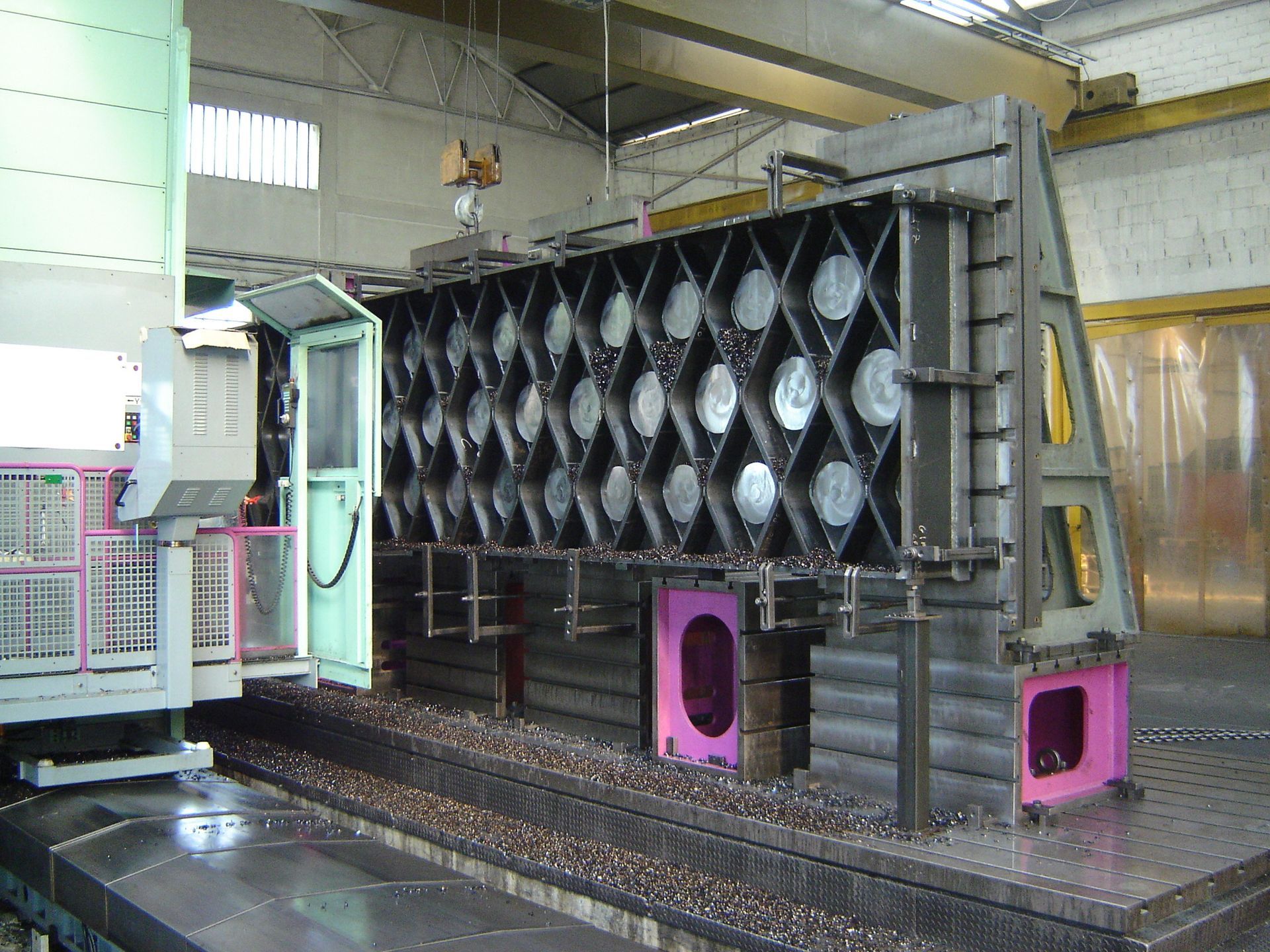 Grandi componenti metallici industriali all'interno di un'officina di fabbrica con una macchina DMG blu sullo sfondo.