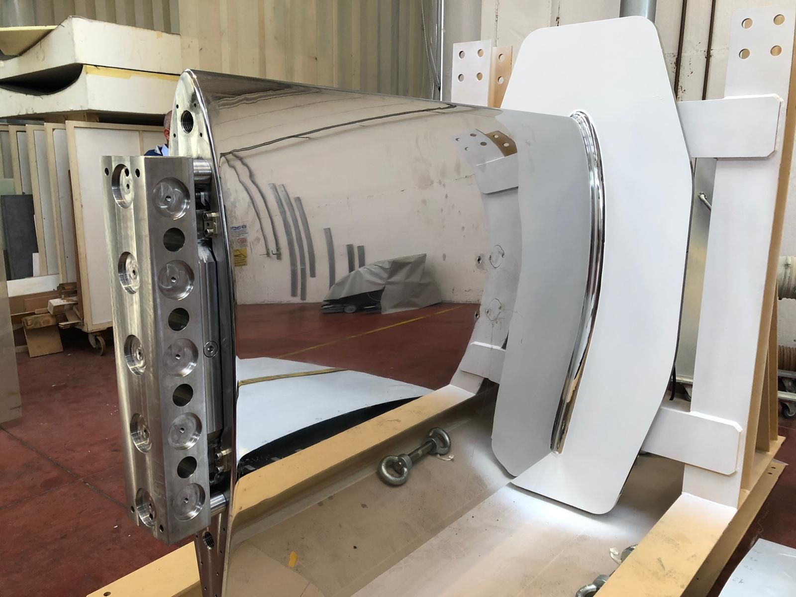Grande sezione metallica dello scafo di una nave in un magazzino, con uno yacht sullo sfondo.