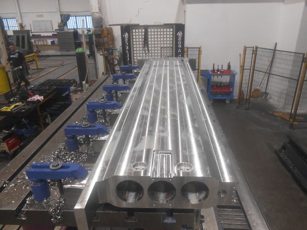 Ampia sezione di scafo in acciaio all'interno di un'officina industriale, con uno yacht sullo sfondo.