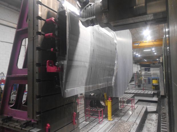 Cantiere navale industriale con componenti in acciaio, gru e un grande scafo di nave davanti a un magazzino blu.