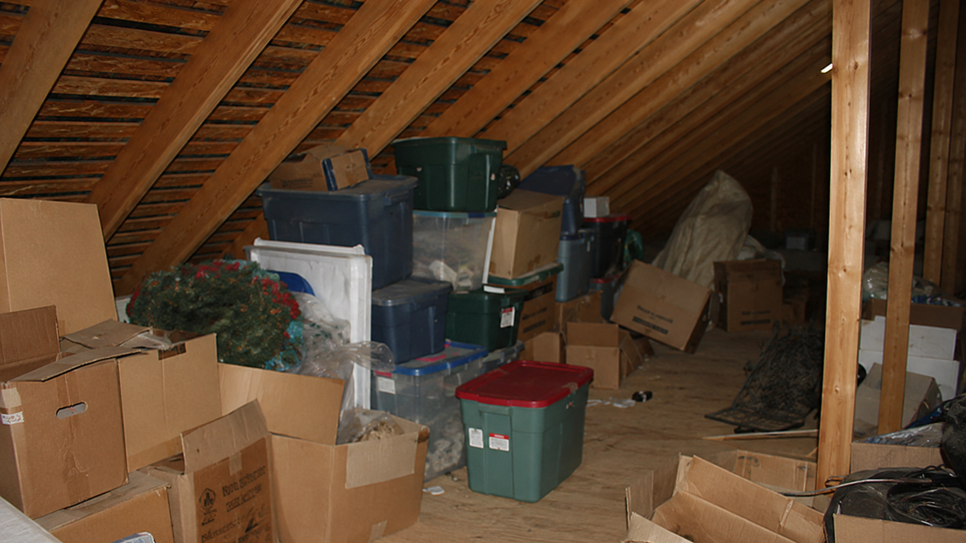 Cluttered attic with stacked boxes, plastic bins, and miscellaneous stored items under a sloped wooden roof
