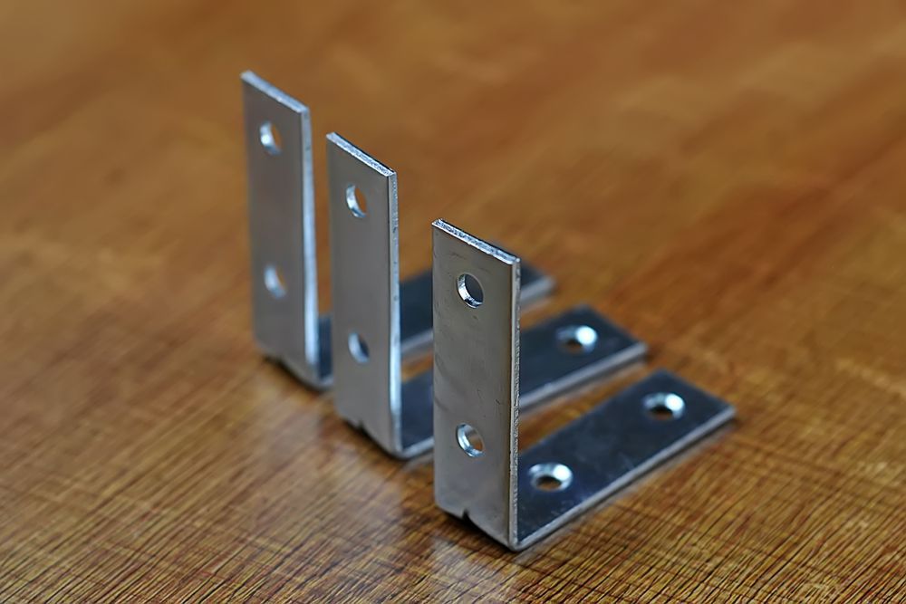 Four silver L-shaped metal brackets with screw holes, arranged on a wood surface — CQR Locksmiths In Moree, NSW