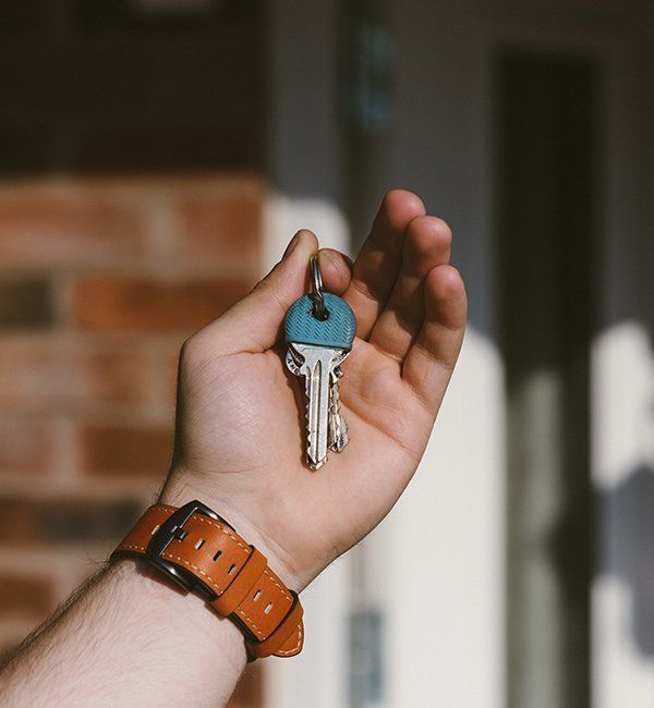Hand Holding Keys In Front Of A Brick Building and White Door. Brown Watch On Wrist — CQR Locksmiths In Moree, NSW