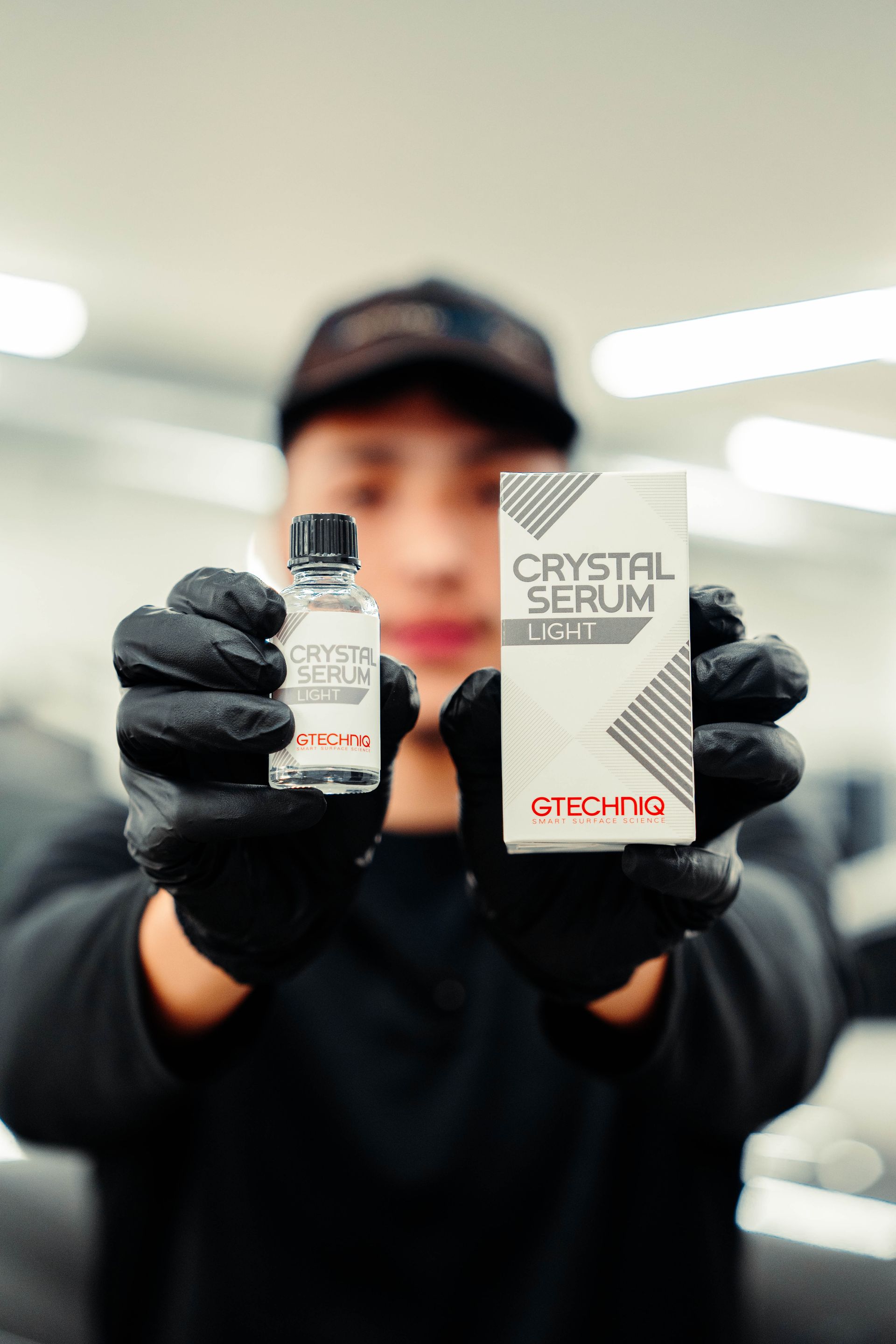 Person in black gloves holding a small bottle and a boxed crystal serum in a blurred indoor setting