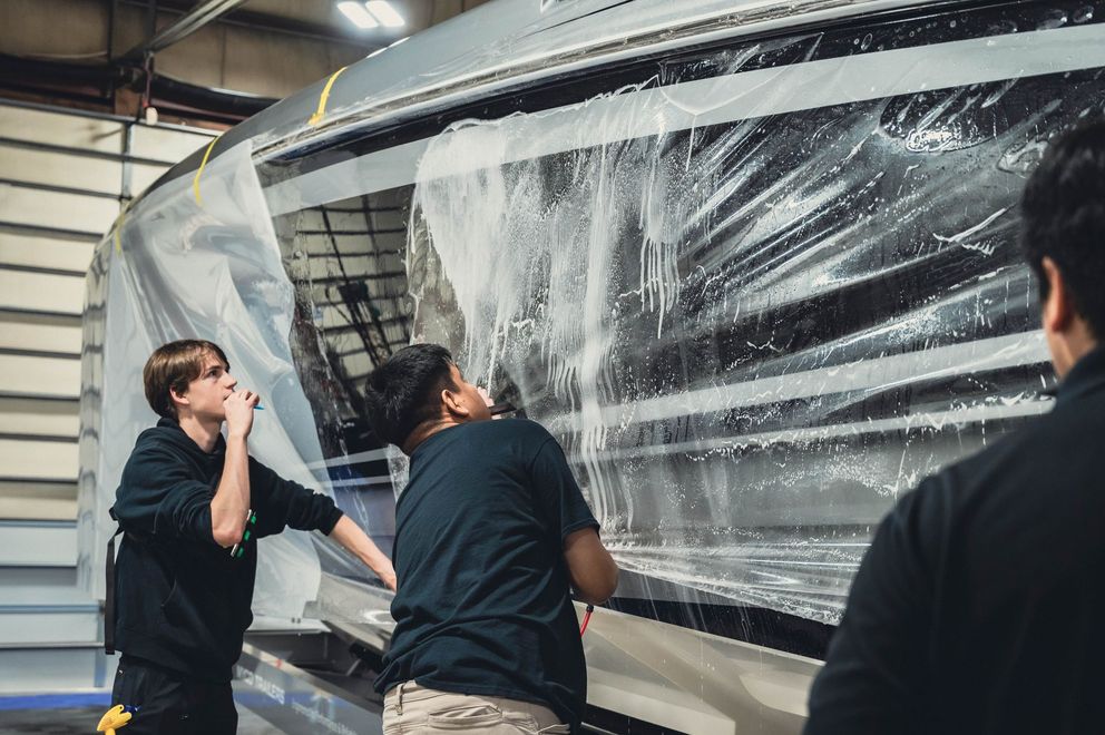 Three people wrapping a large boat in clear plastic inside a workshop