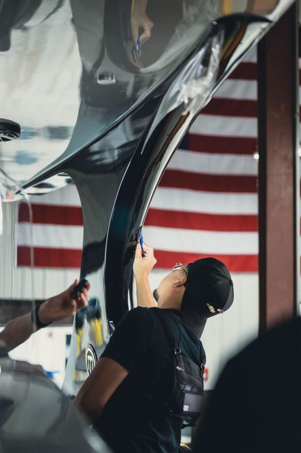 Person holds up a shiny aircraft fuselage section in a workshop with a U.S. flag in the background
