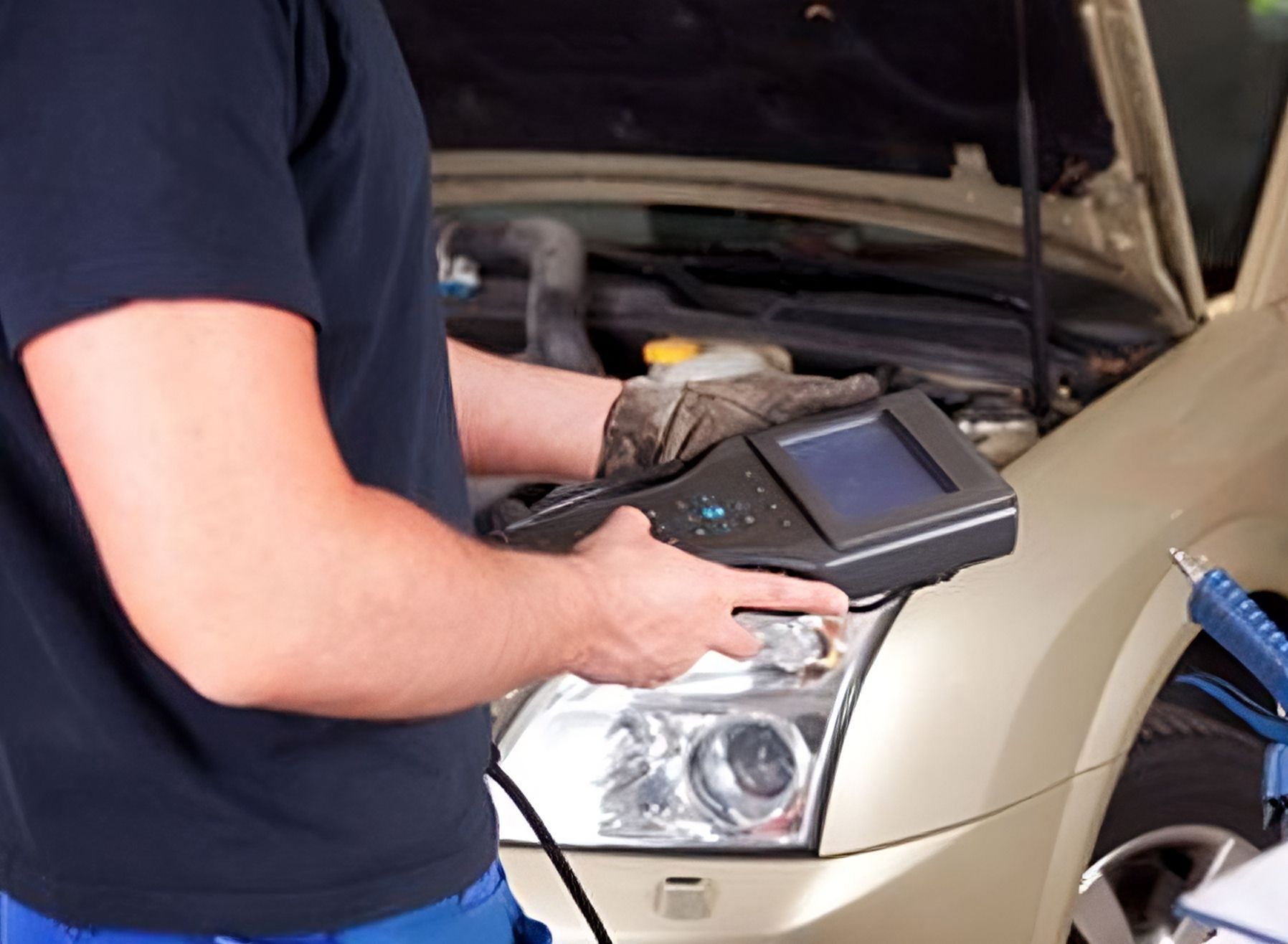 A man is holding a device in front of a car with the hood open.