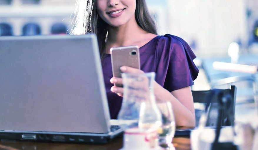 A woman is sitting at a table with a laptop and a cell phone.