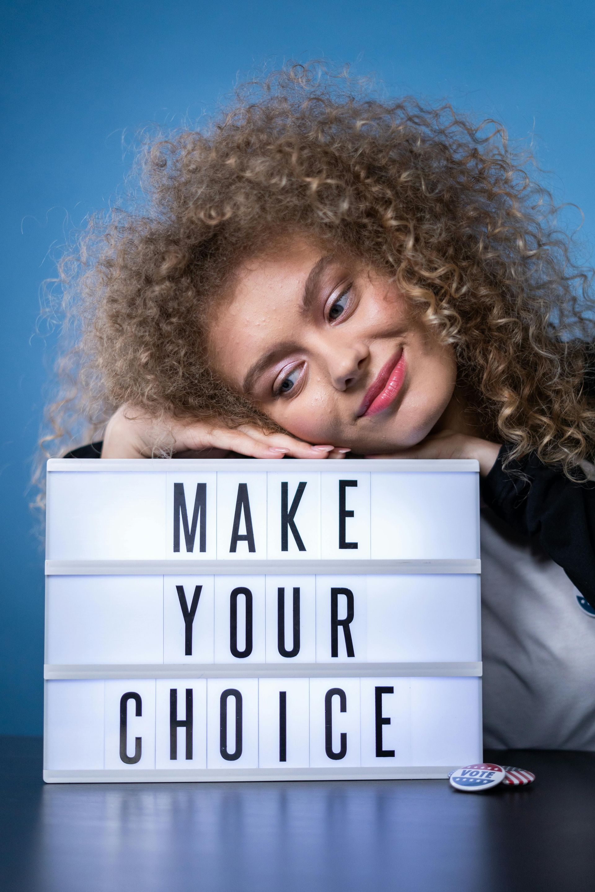 A white lady with curly hair resting her head on a sign that says make your choice.