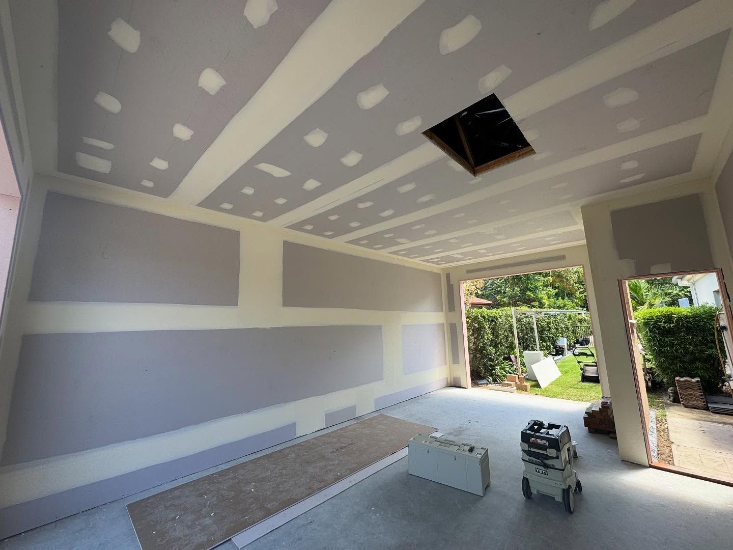 Unfinished room with drywall patching, a skylight opening, and a view to a garden through an open doorway.