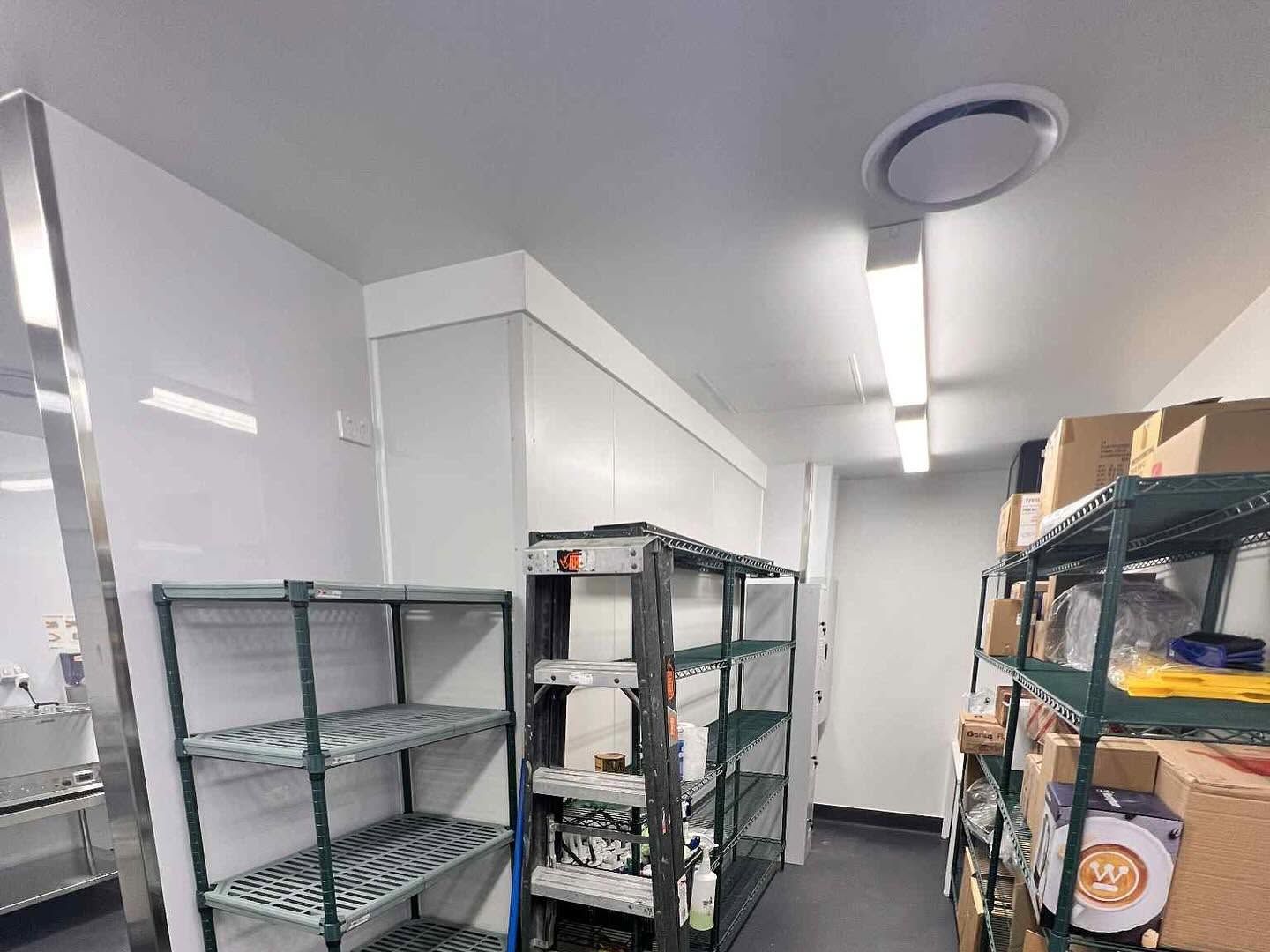 Storage room with empty wire racks, white cabinets, and boxes in a narrow aisle under fluorescent lights