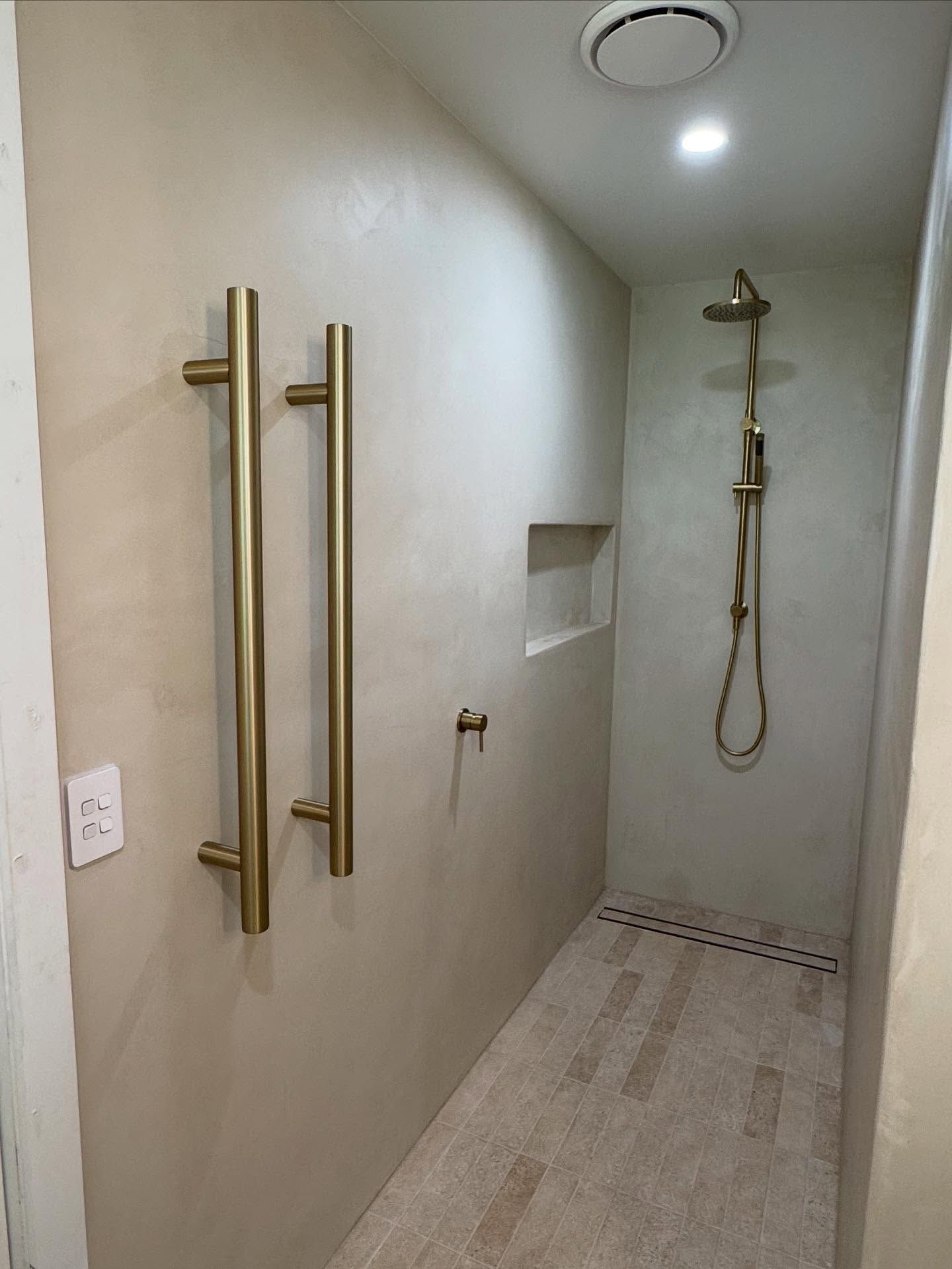 Accessible shower with gold grab bars and handheld showerhead in a narrow bathroom stall