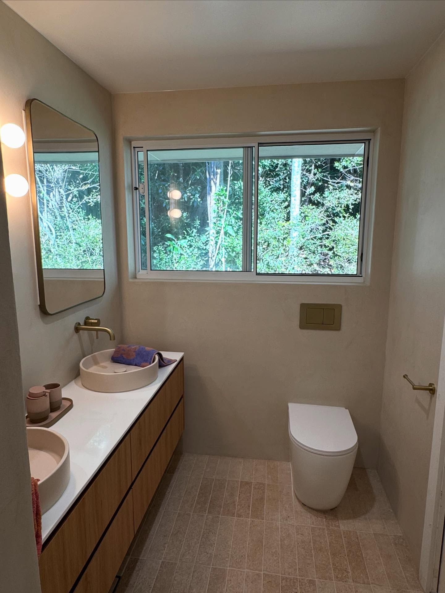 Modern bathroom with double sink vanity, mirror lights, window, and toilet; neutral tones and wood accents