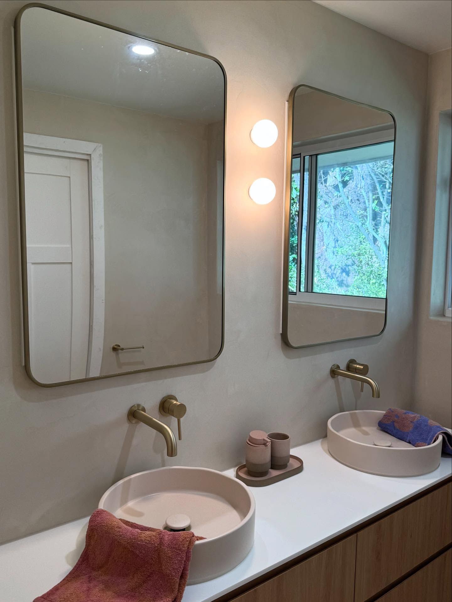 Bathroom vanity with two round sinks, brass faucets, mirrors, and a window view
