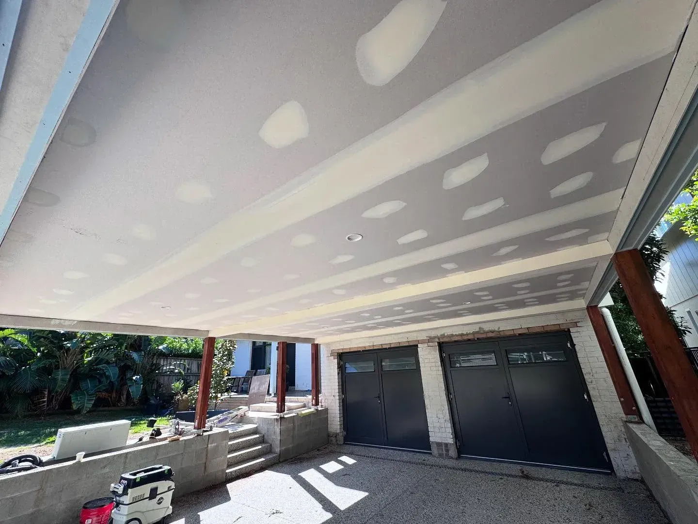 Covered driveway under construction with unfinished drywall ceiling and black garage doors beside a concrete entryway