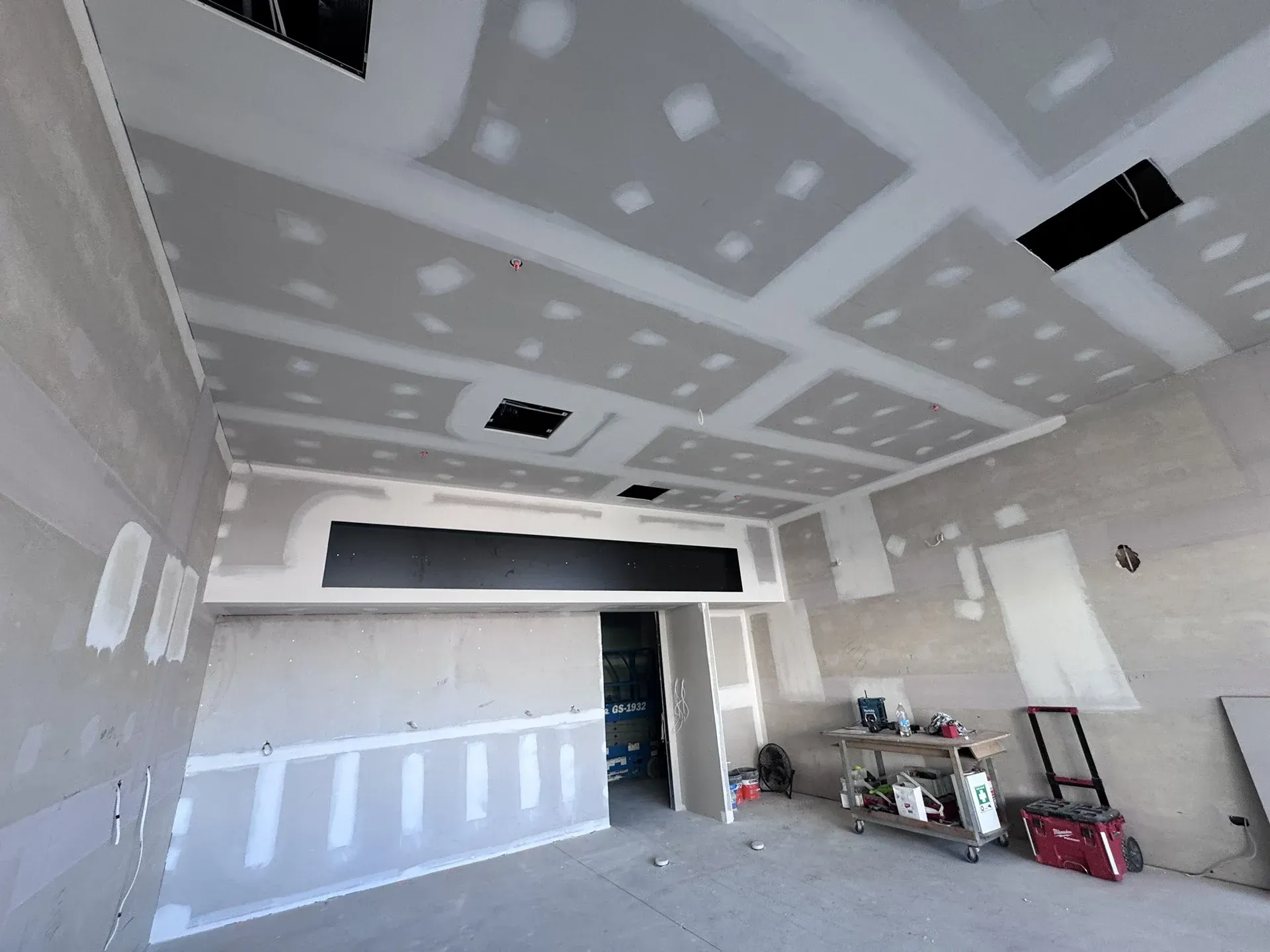 Unfinished room with drywall patches on ceiling and walls, tools and supplies along the right side.