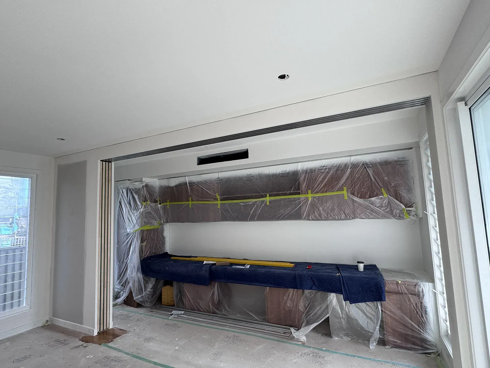 Living room under renovation, with plastic-covered wall opening and blue padding across the center.