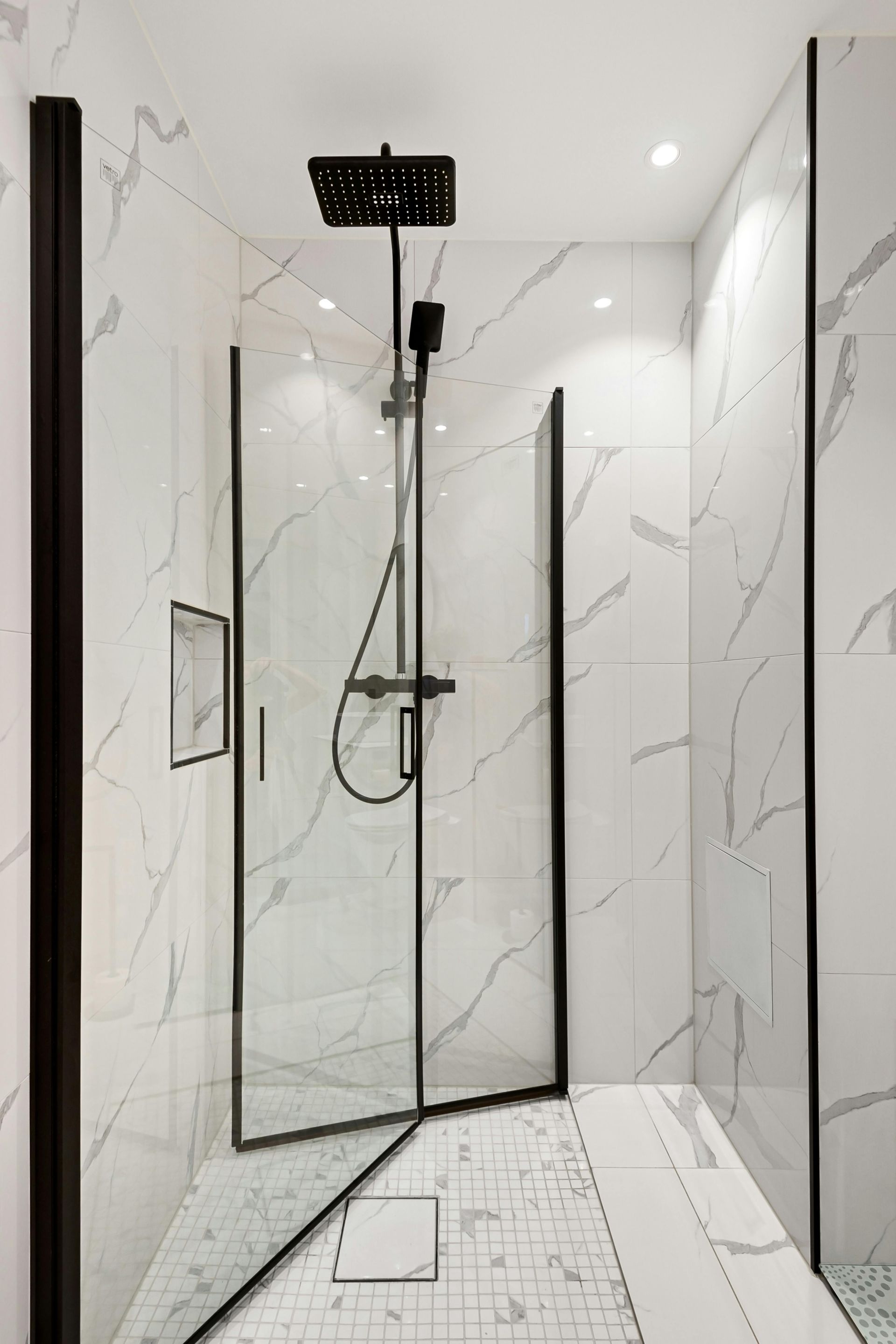 A bathroom with white tiles and a walk in shower with a black shower head.